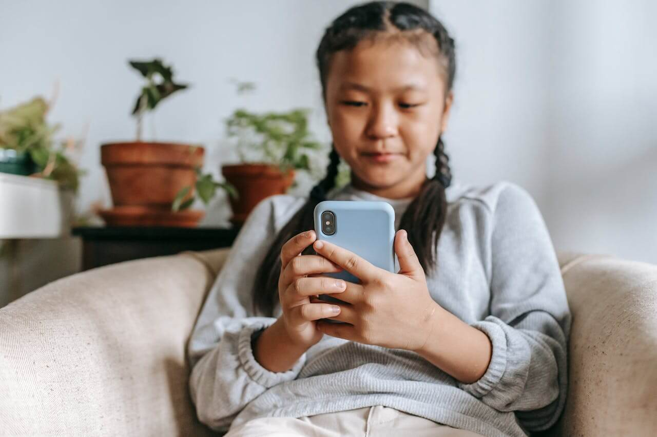 A young girl with braided hair sitting in an armchair while looking at a smartphone.