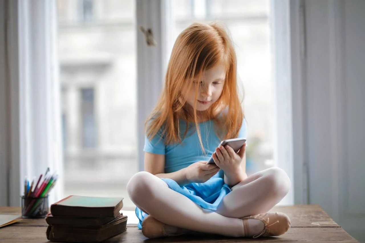 A young red-haired girl in a blue dress sitting cross-legged while looking at a smartphone.