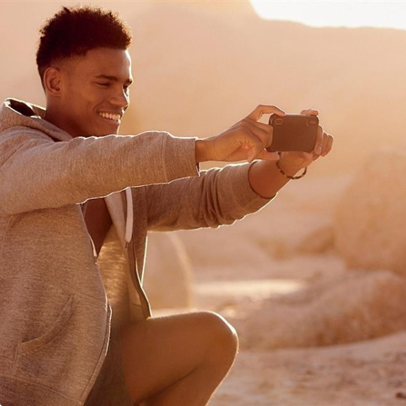 Smiling young man kneeling outdoors in warm sunlight, taking a photo horizontally with his smartphone.